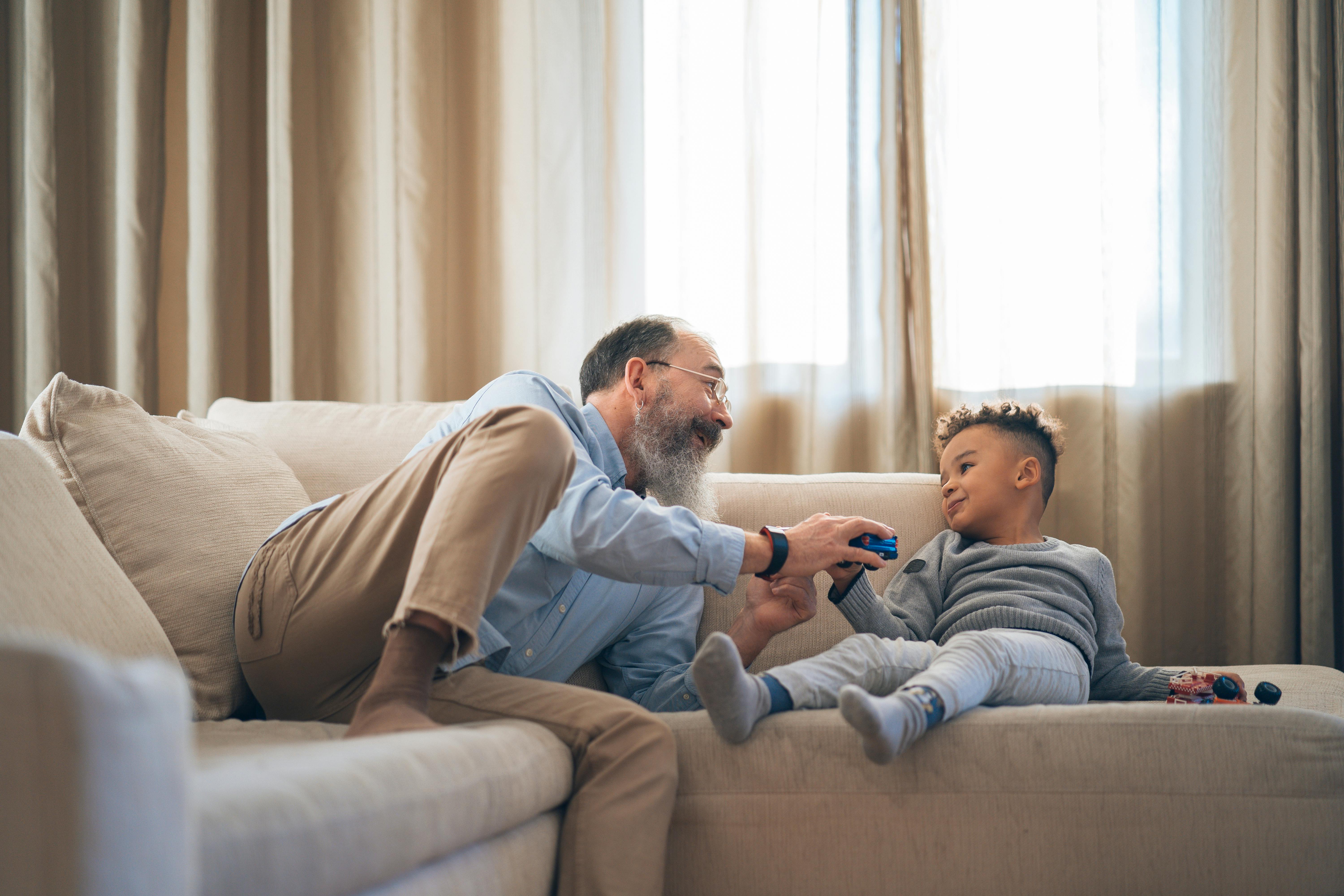 Smiling grandfather playing with his young grandson while sitting together on a sofa at home, symbolising family bonds and generational care.